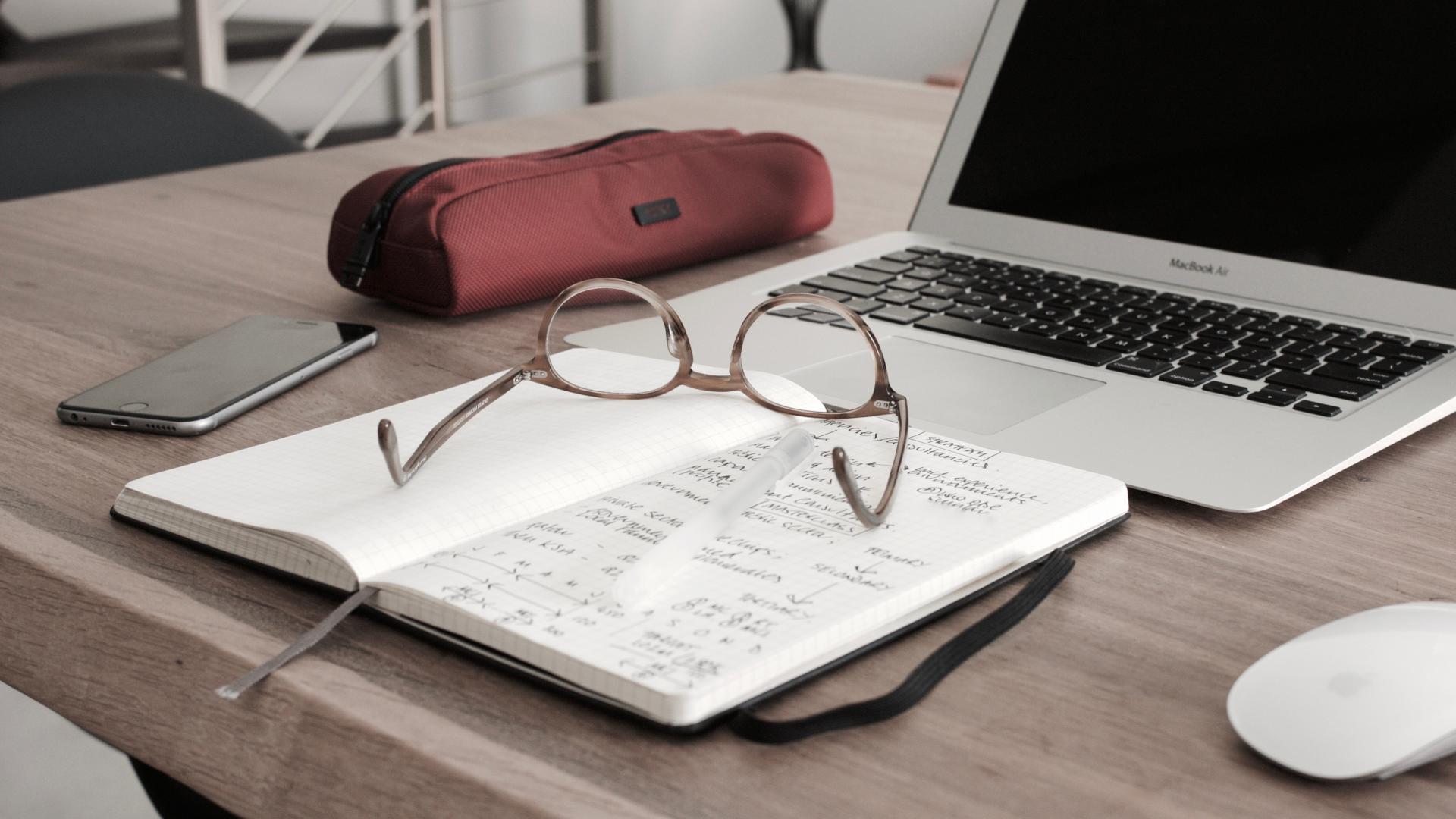 A table with a laptop computer, mobile phone, notebook and glasses.