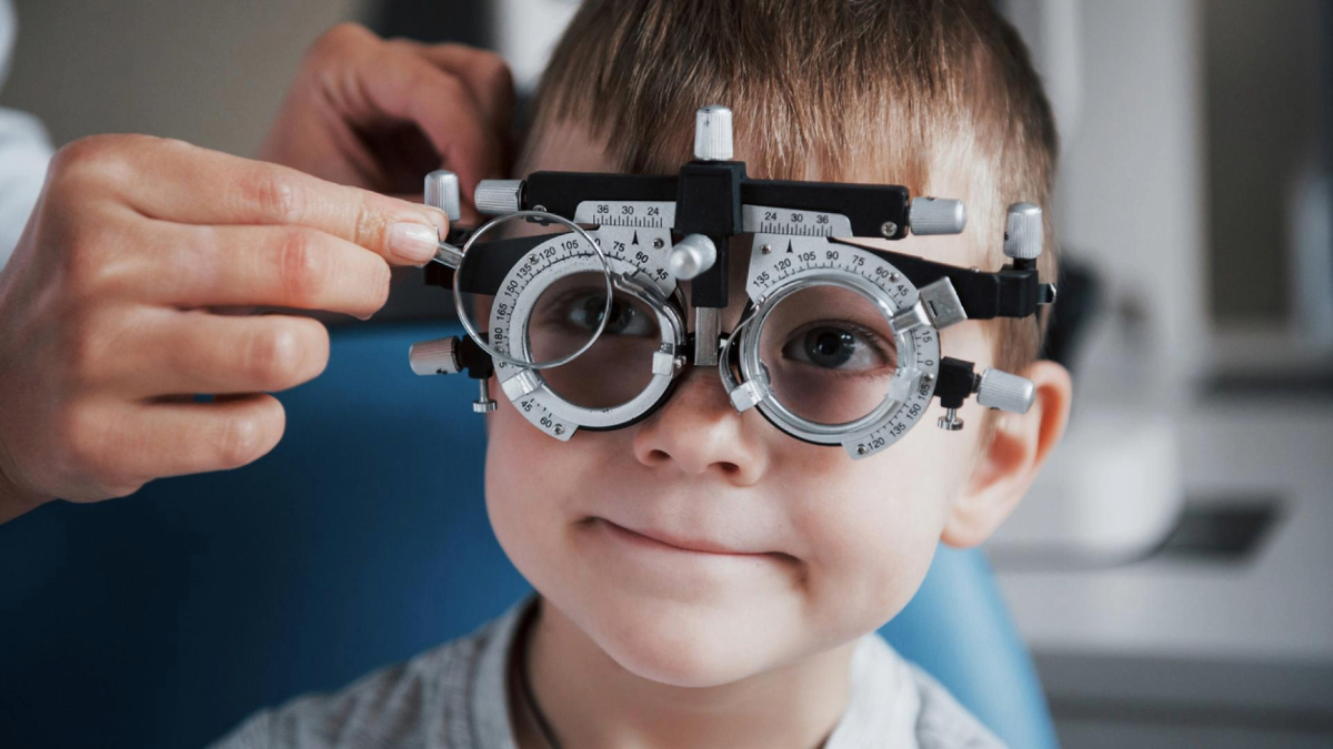 A child is smiling while undergoing an eye exam with a phoropter to assess vision.