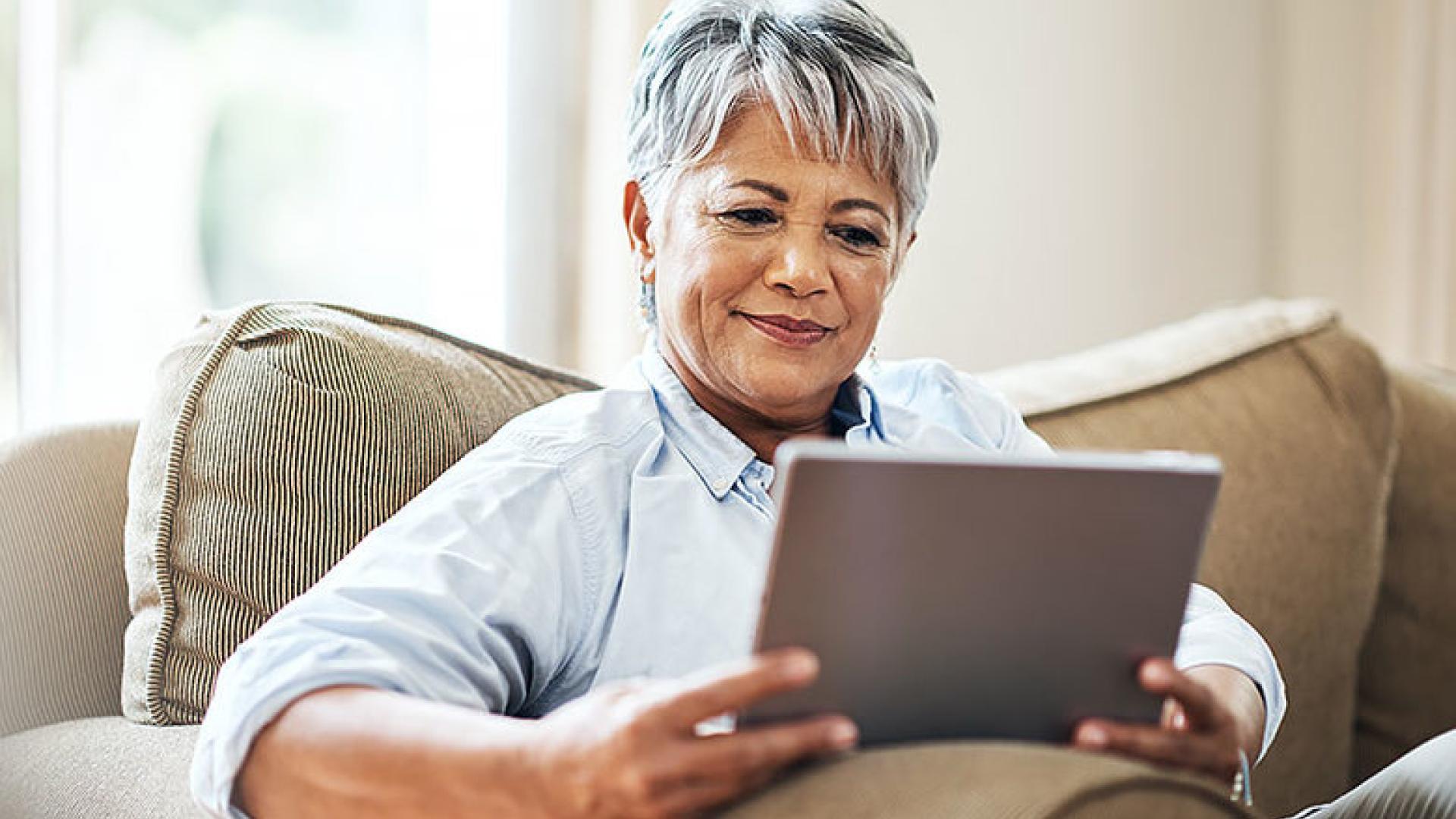 Senior adult sitting on a couch and looking at a digital tablet device.