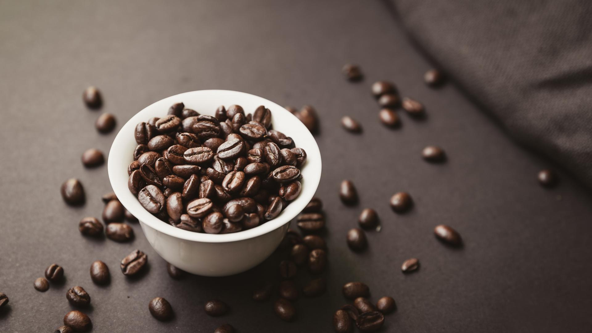 a white bowl full of roasted coffee beans sits on a dark table with more beans scattered on its surface