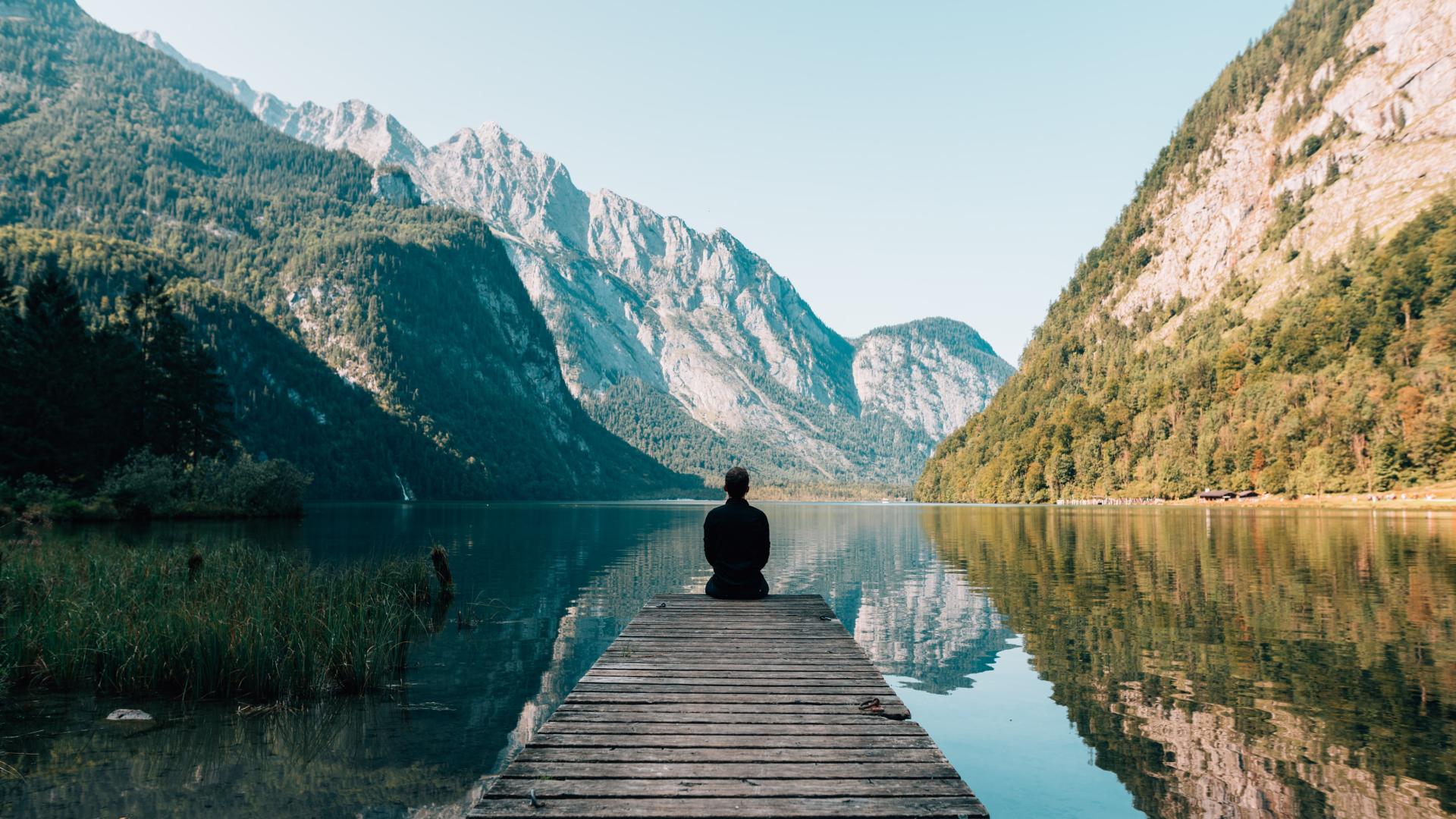 Someone sits at the end of a dock overlooking a placid mountain valley lake.