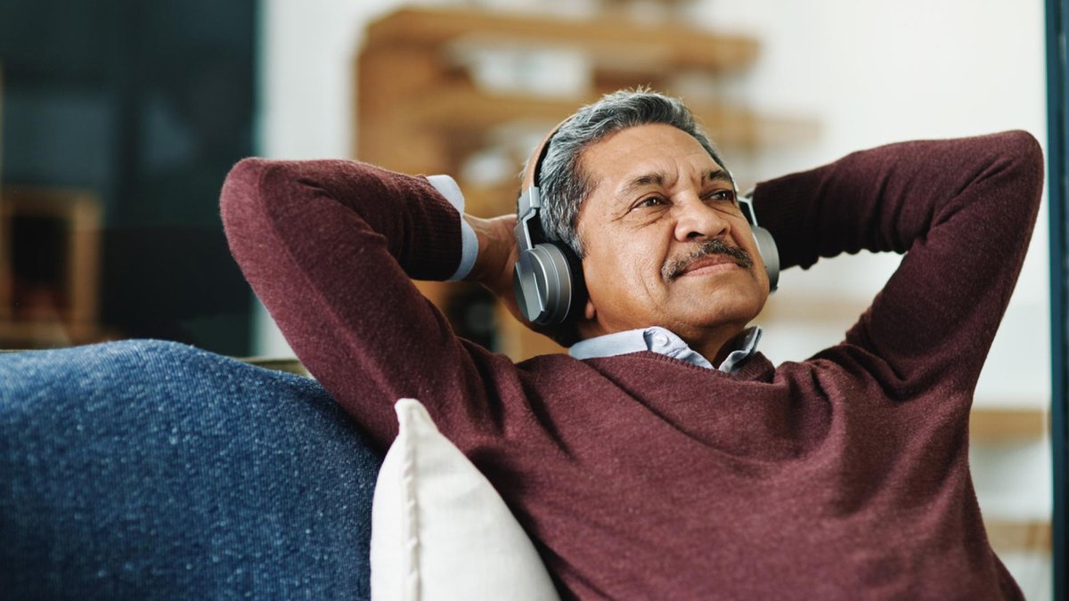 An older man relaxes on a sofa, wearing headphones and leaning back with his hands behind his head.