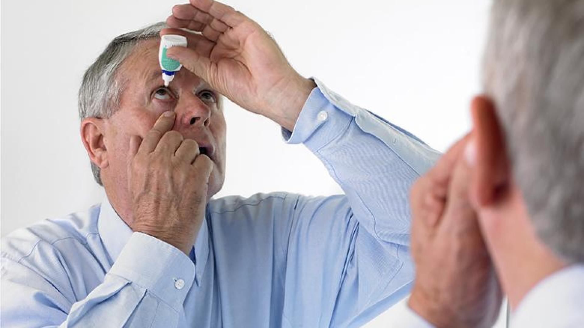 A senior man applies eyedrops in front of a mirror.