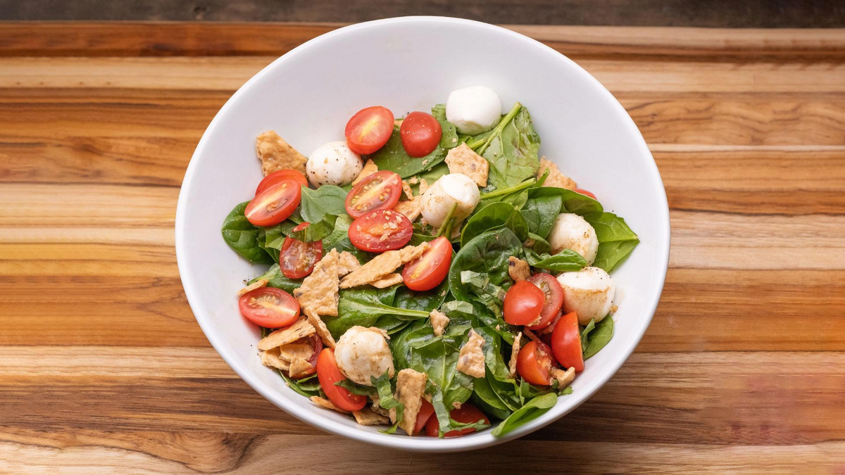 A bowl of salad containing spinach leaves, halved cherry tomatoes, mozzarella balls, and crispy bread pieces, served on a wooden surface.