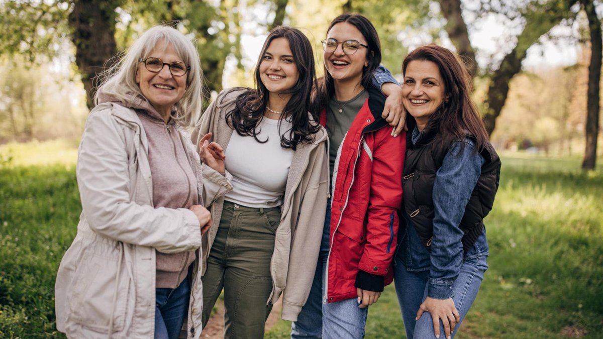 A joyful, multigenerational group of women, including an elderly woman, stand outdoors in a park, smiling and embracing each other.