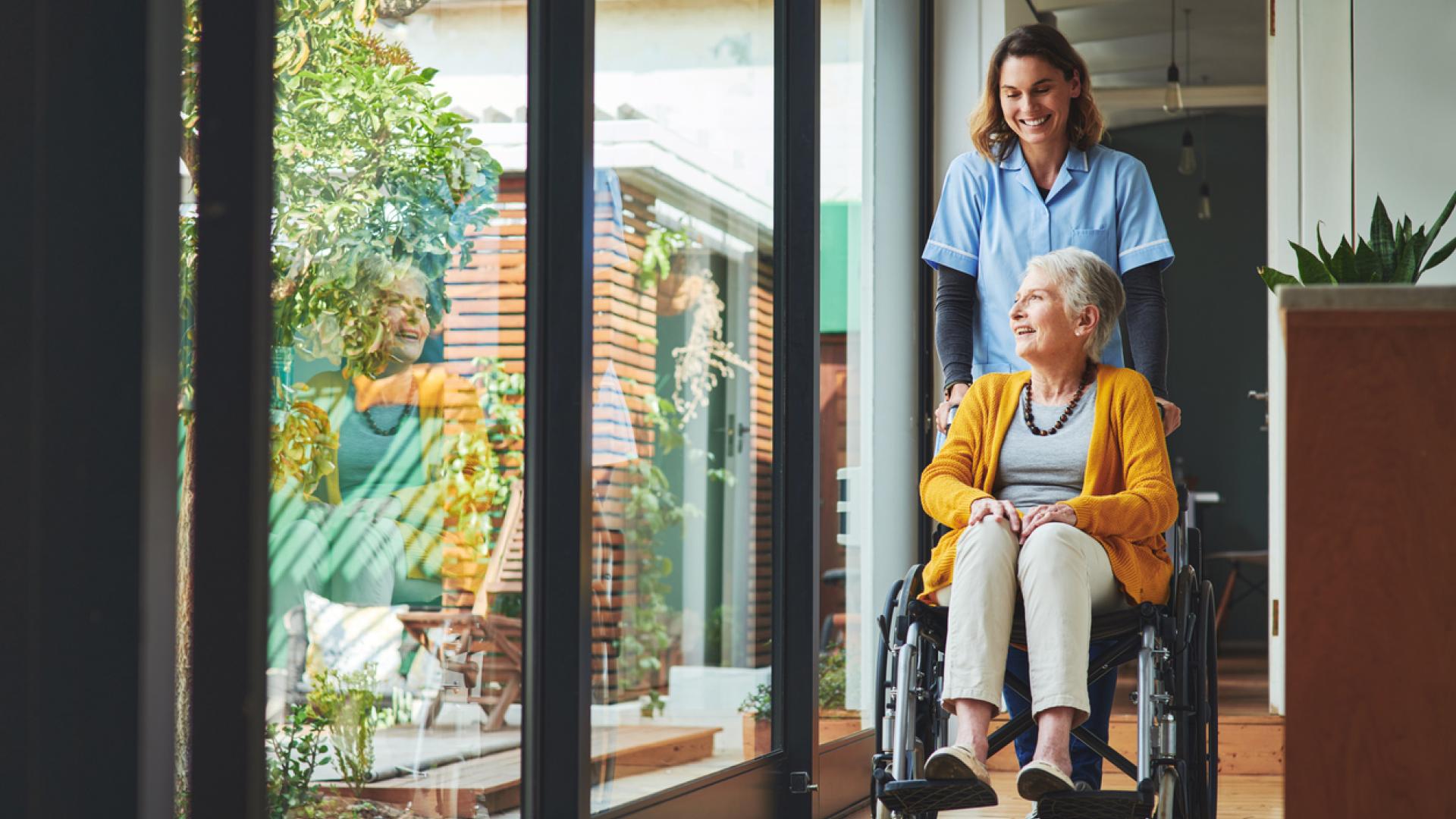 An elderly woman in a wheelchair stares out a floor-to-ceiling window while a young woman in nurse's scrubs pushes her chair down a hallway.