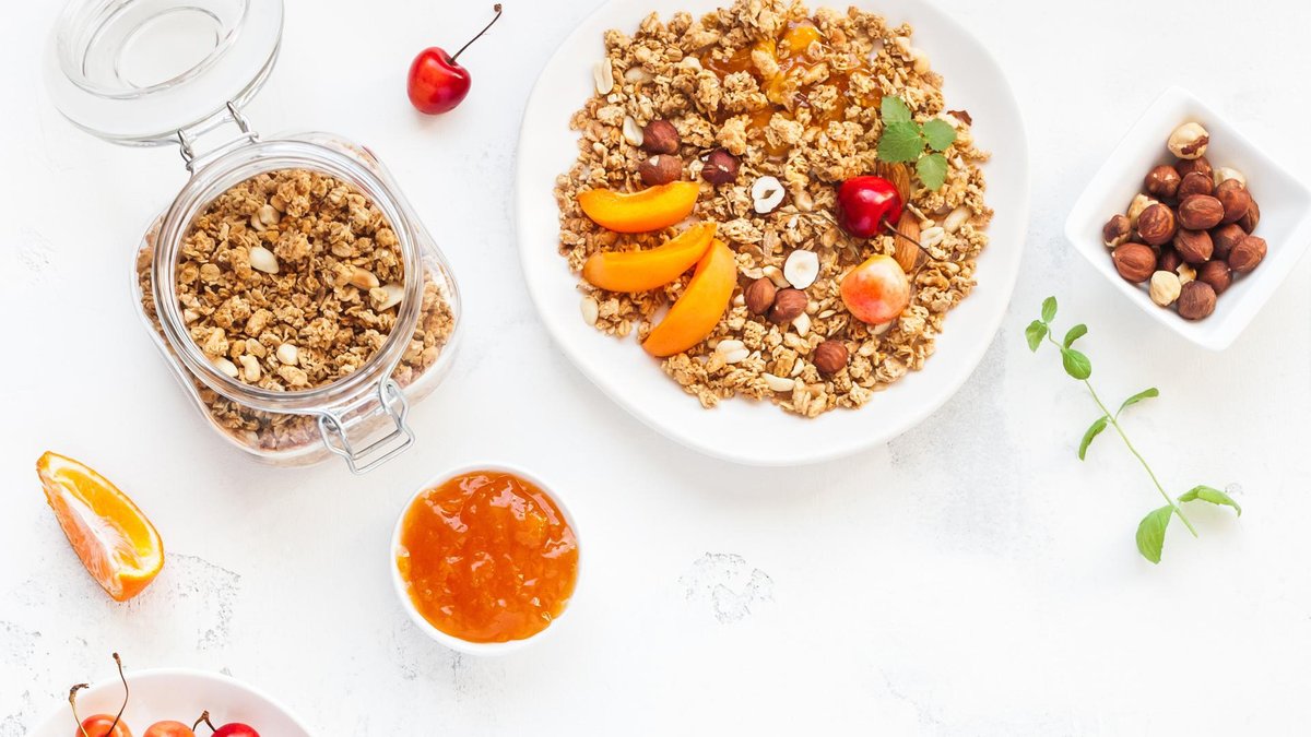 A white bowl filled with granola topped with slices of fresh peach, cherries, and hazelnuts. The bowl is surrounded by additional healthy ingredients like a jar of granola, cherries, and a small bowl of orange jam.