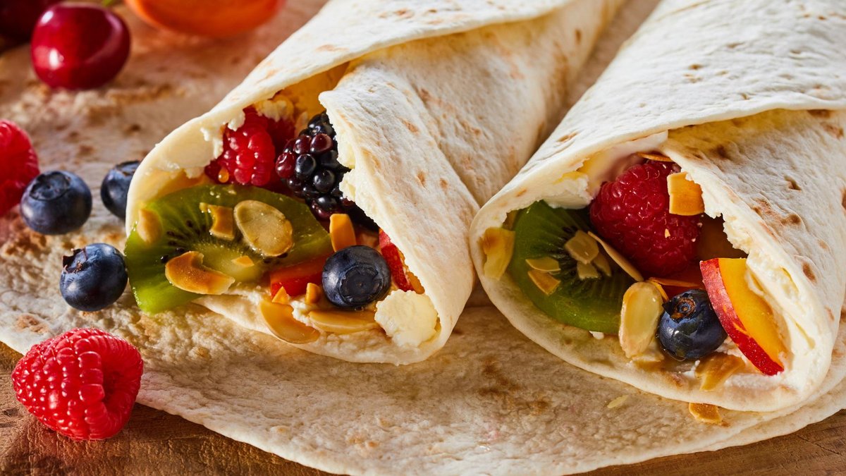 Close-up of two rolled tortillas filled with fresh fruit, including kiwi, berries, and almonds, served on a wooden board.