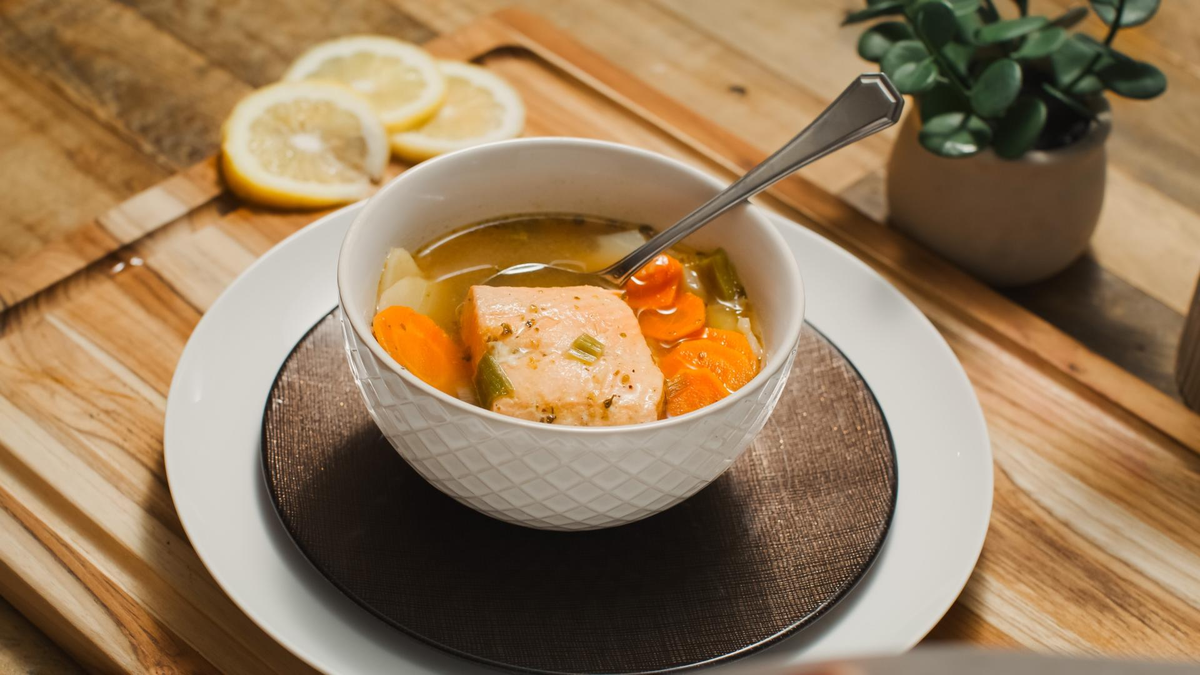 A bowl of warm soup containing salmon, carrots, and vegetables, served in a white ceramic bowl on a wooden placemat. Thin lemon slices are placed on the table beside the bowl, contributing to a cozy and appetizing presentation.