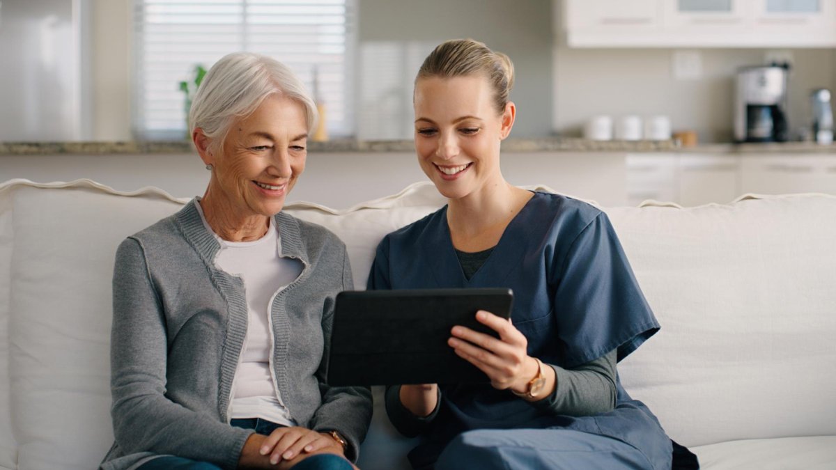 A caregiver in medical scrubs sits on a couch with a senior woman, both smiling while looking at a tablet.