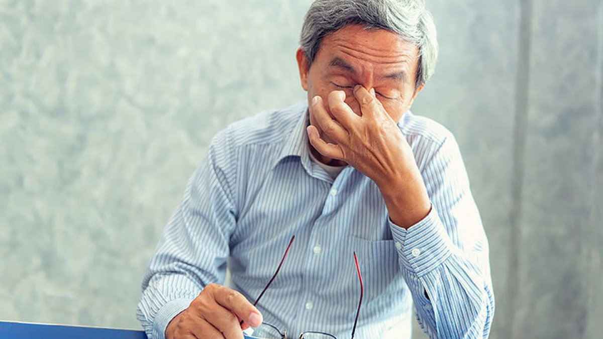 An elderly man wearing a striped shirt rubs his eyes while holding his glasses, appearing to be in discomfort, possibly due to a headache or eye strain. The background is out of focus, drawing attention to the man's expression.