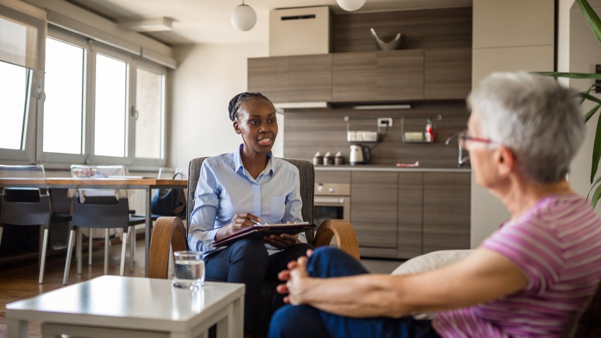 A healthcare professional seated in a modern home, discussing care with an elderly woman. The scene reflects a warm, comfortable environment with a focus on health and support.