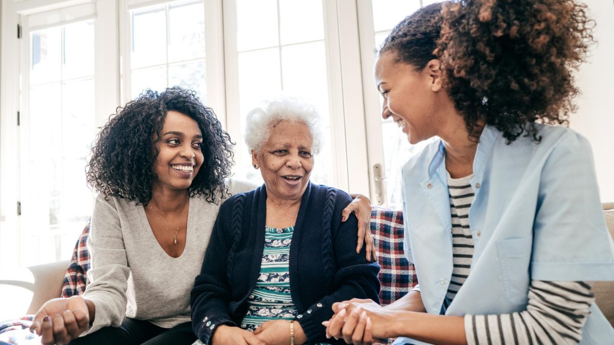 A senior woman sits on a couch between a younger woman and a caregiver, all smiling and talking together. The caregiver is wearing a light blue uniform, and the scene conveys warmth and support in a caregiving setting.