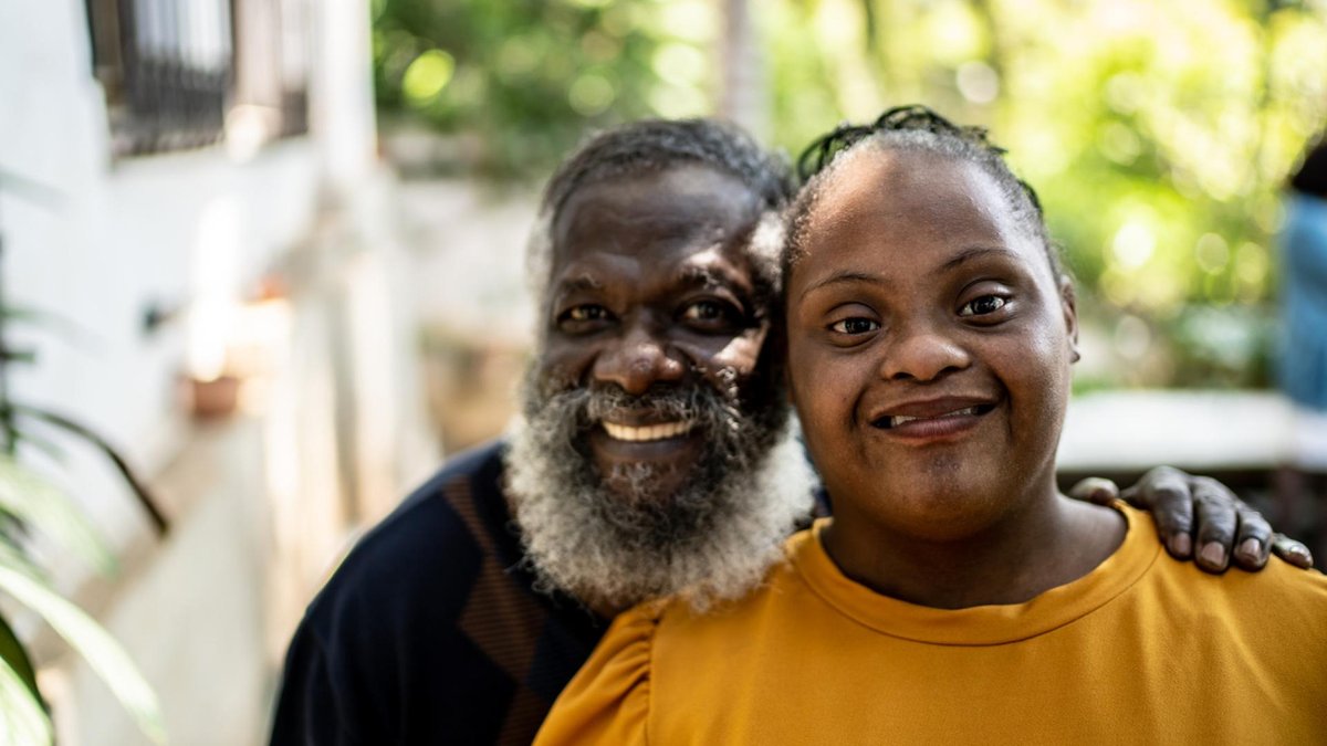 A father and his daughter, who has Down syndrome, are outdoors. They both smile warmly at the camera, with the father affectionately placing his arm around his daughter in a lush, green setting.