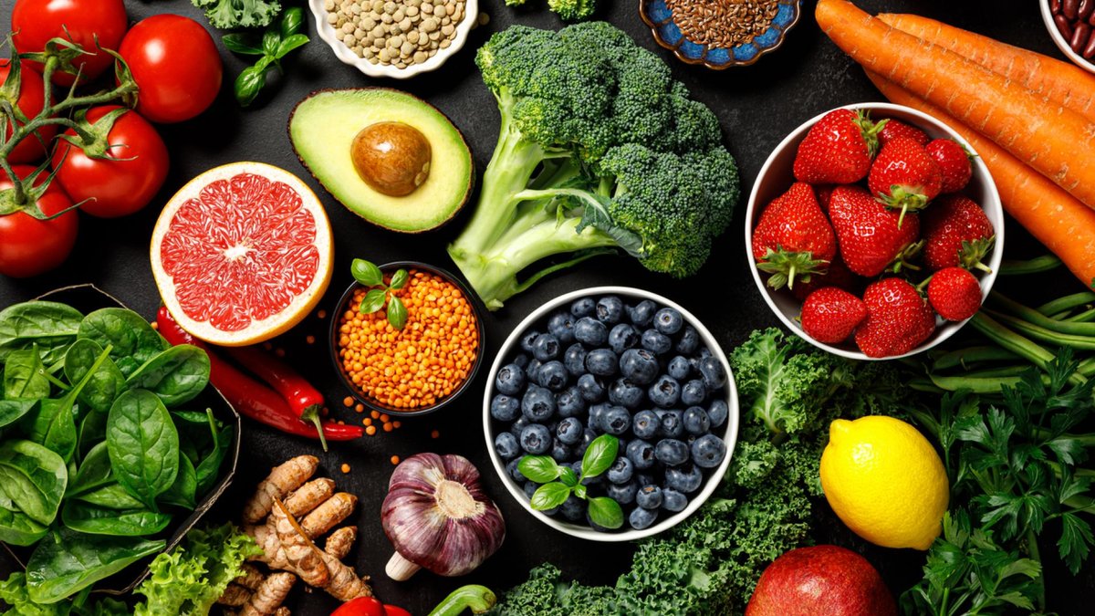 A vibrant assortment of fruits, vegetables, and healthy ingredients spread across a dark background. Items include tomatoes, avocado, broccoli, blueberries, strawberries, grapefruit, spinach, and more, showcasing a colorful variety of fresh produce.