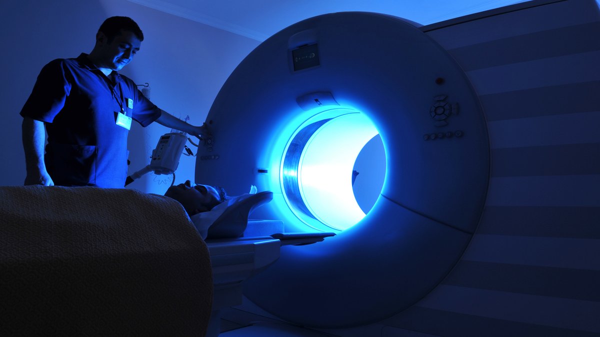 A patient undergoing an MRI scan in a hospital setting. The room is dimly lit with a blue glow, and a healthcare professional is standing beside the MRI machine, overseeing the procedure.