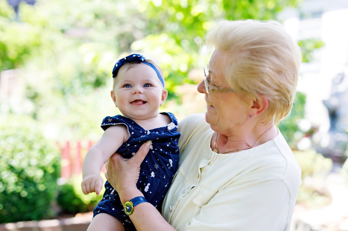 A joyful grandmother holding her smiling baby granddaughter in an outdoor setting with greenery in the background.