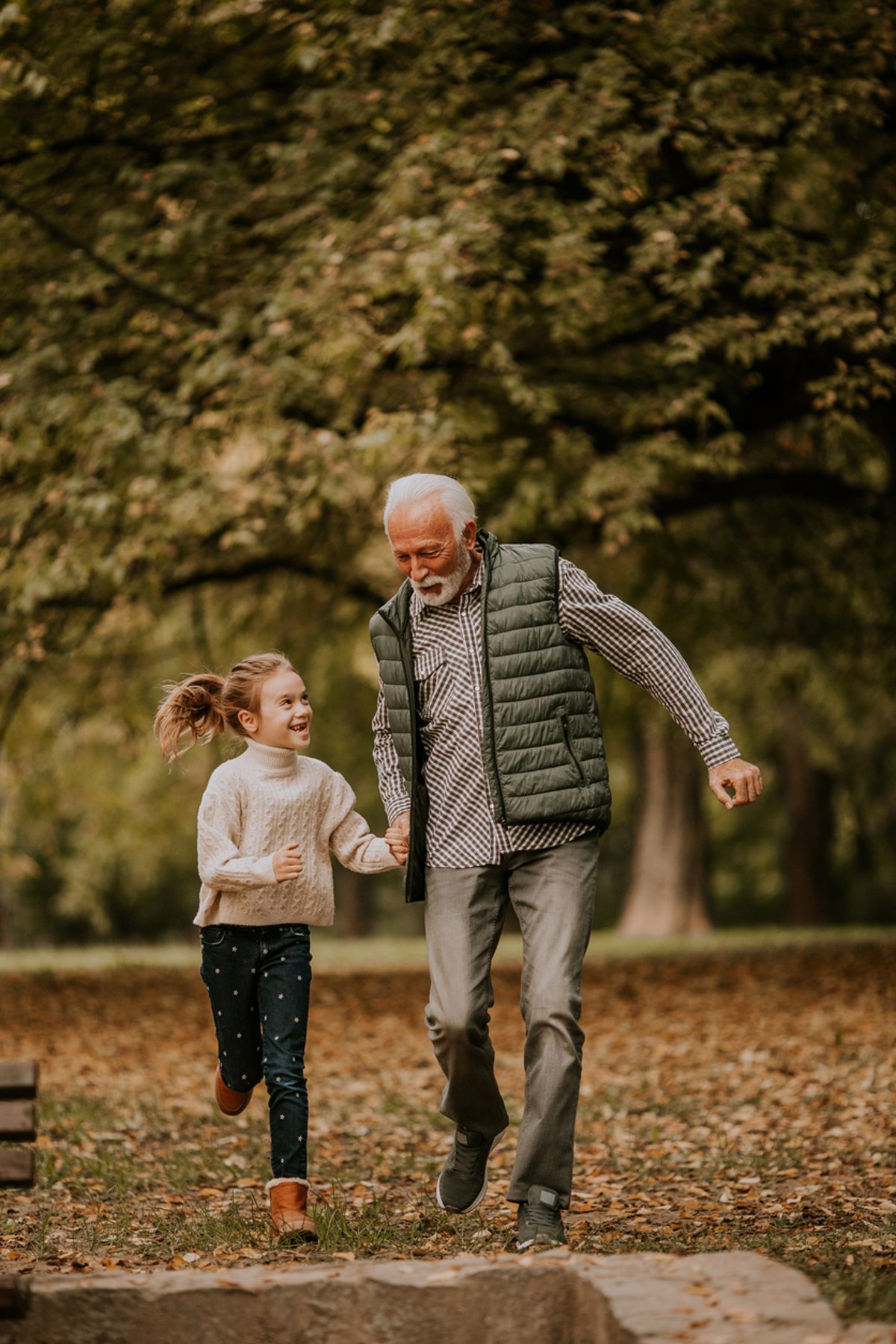 An elderly man and a young girl joyfully running together through a park on an autumn day. The man holds the girl's hand as they smile, surrounded by trees with fallen leaves covering the ground.