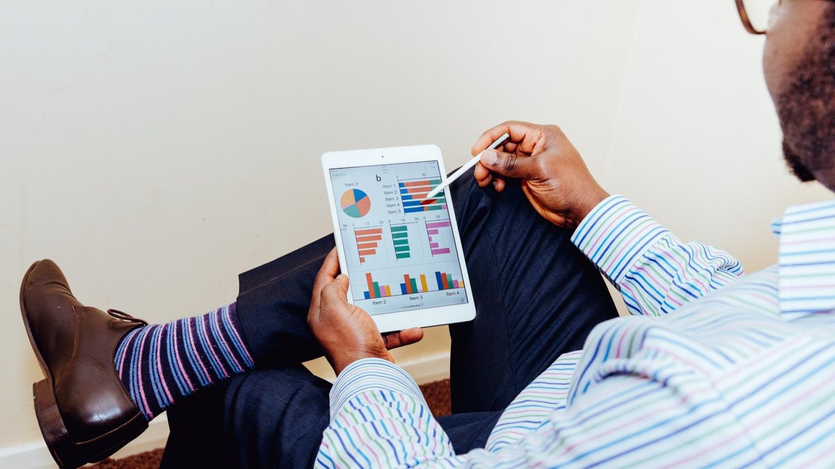 A person holding a tablet displaying various colorful charts and graphs, sitting with one leg crossed and wearing formal shoes and striped socks.