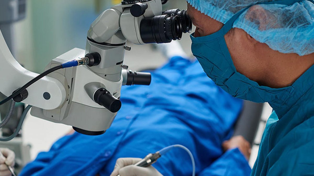 A surgeon performing glaucoma surgery using a microscope. The patient is covered with blue surgical drapes, and the surgeon is wearing a face mask and surgical cap.