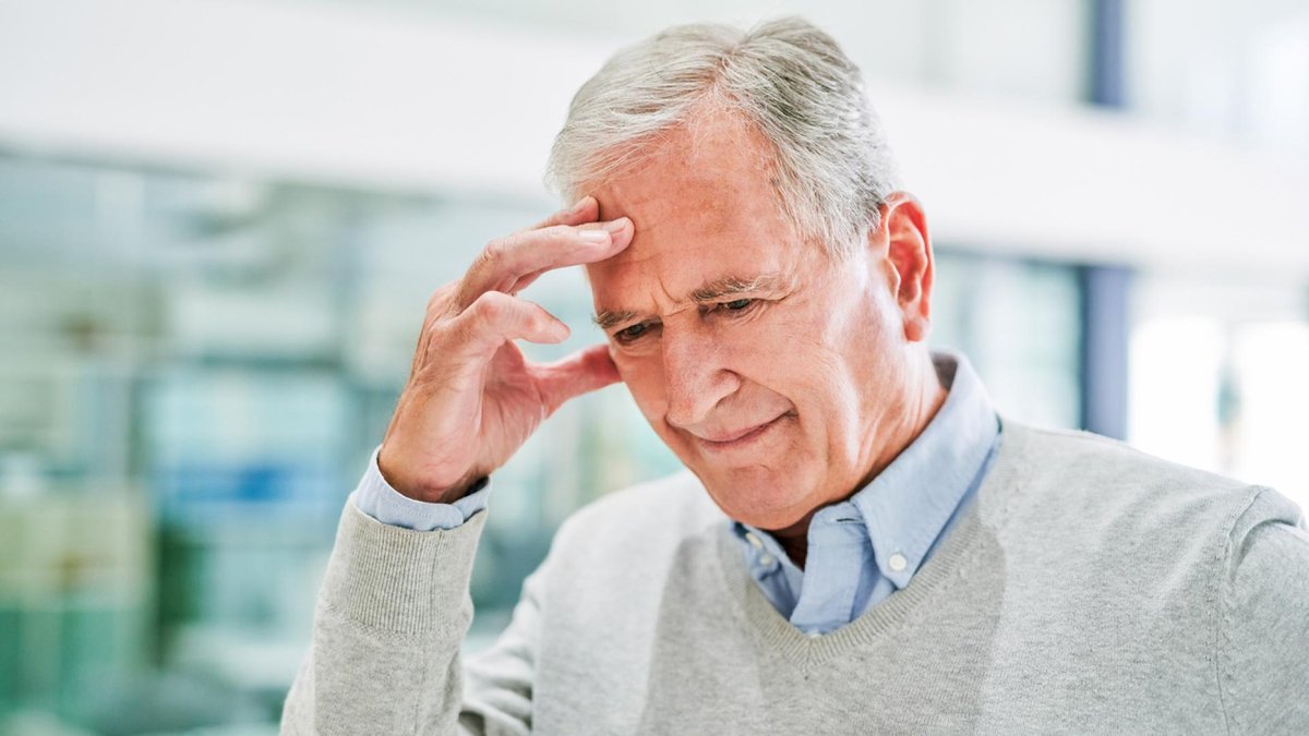 An older man, dressed in a light gray sweater, holds his head with a pained expression.