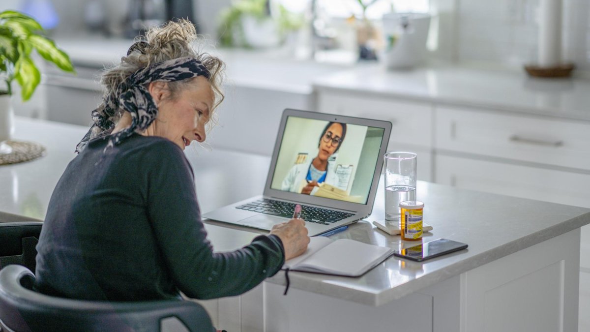 An elderly woman with curly hair, sitting at a kitchen counter, having a video call with a doctor on her laptop. She is taking notes as the doctor, visible on the screen, explains something, with a pill bottle and a glass of water nearby.