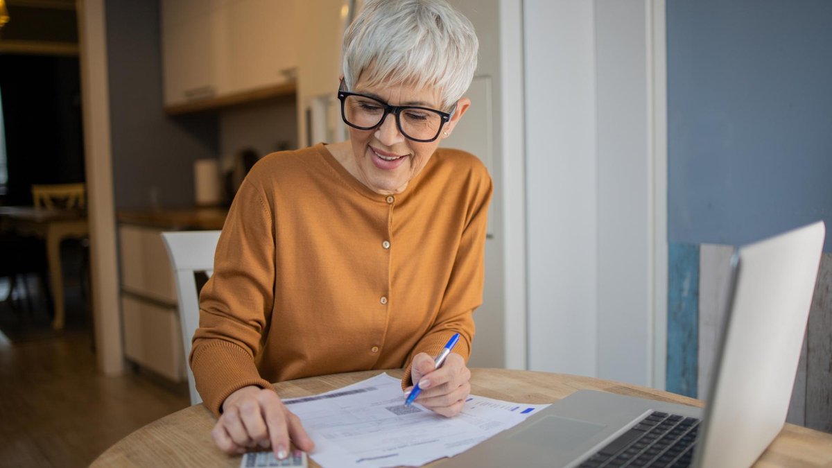 An elderly woman sitting at a table with a calculator and paperwork, reviewing her finances. She appears focused, wearing glasses and working in a well-lit, home-like setting.