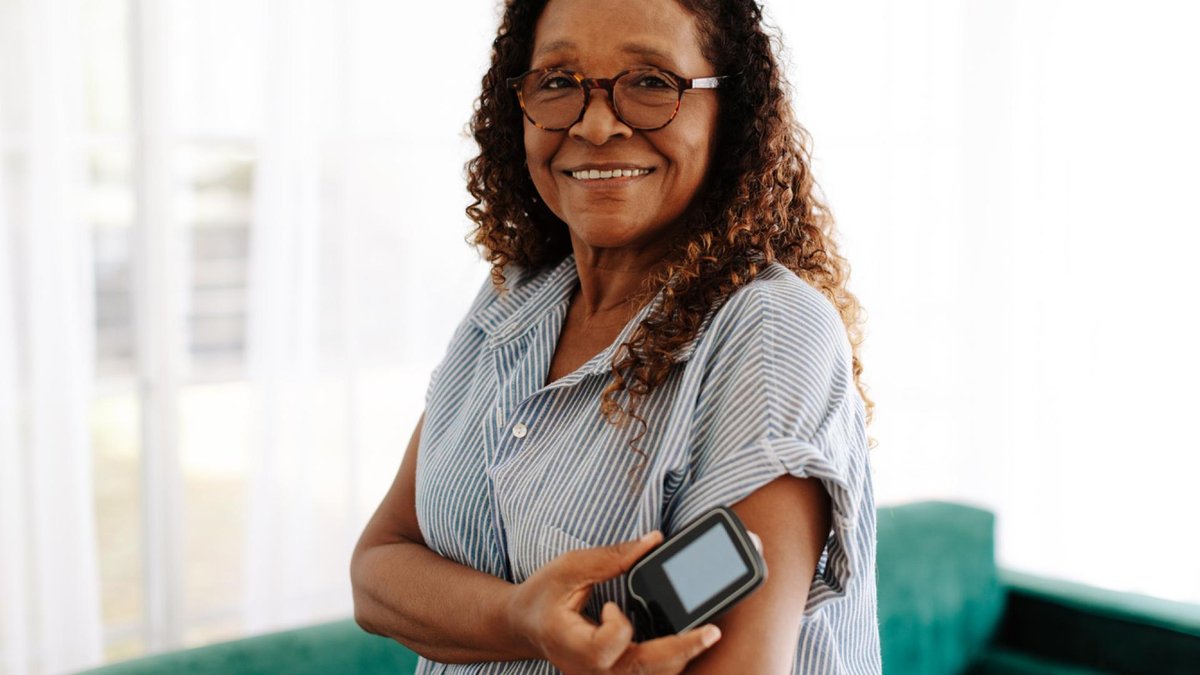 A woman wearing glasses smiles while using a blood glucose monitor to check her blood sugar levels. She is standing in a bright room, appearing confident and comfortable with her health routine.