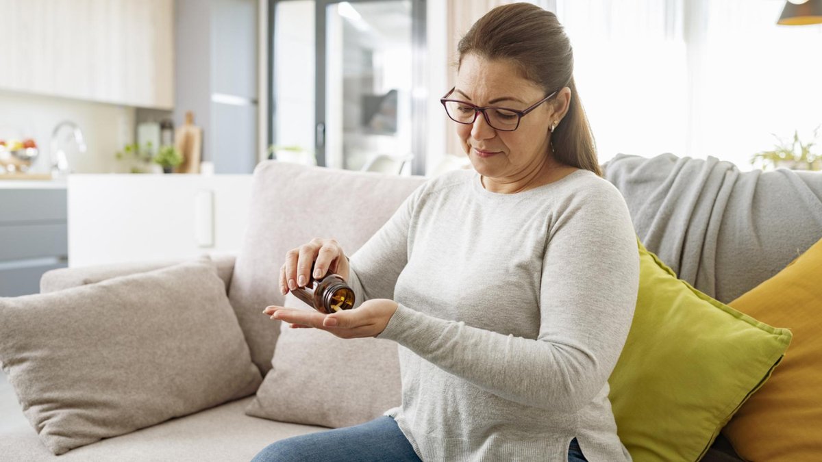 A woman sitting on a couch, carefully pouring pills from a bottle into her hand. She appears thoughtful and focused in her comfortable home environment.