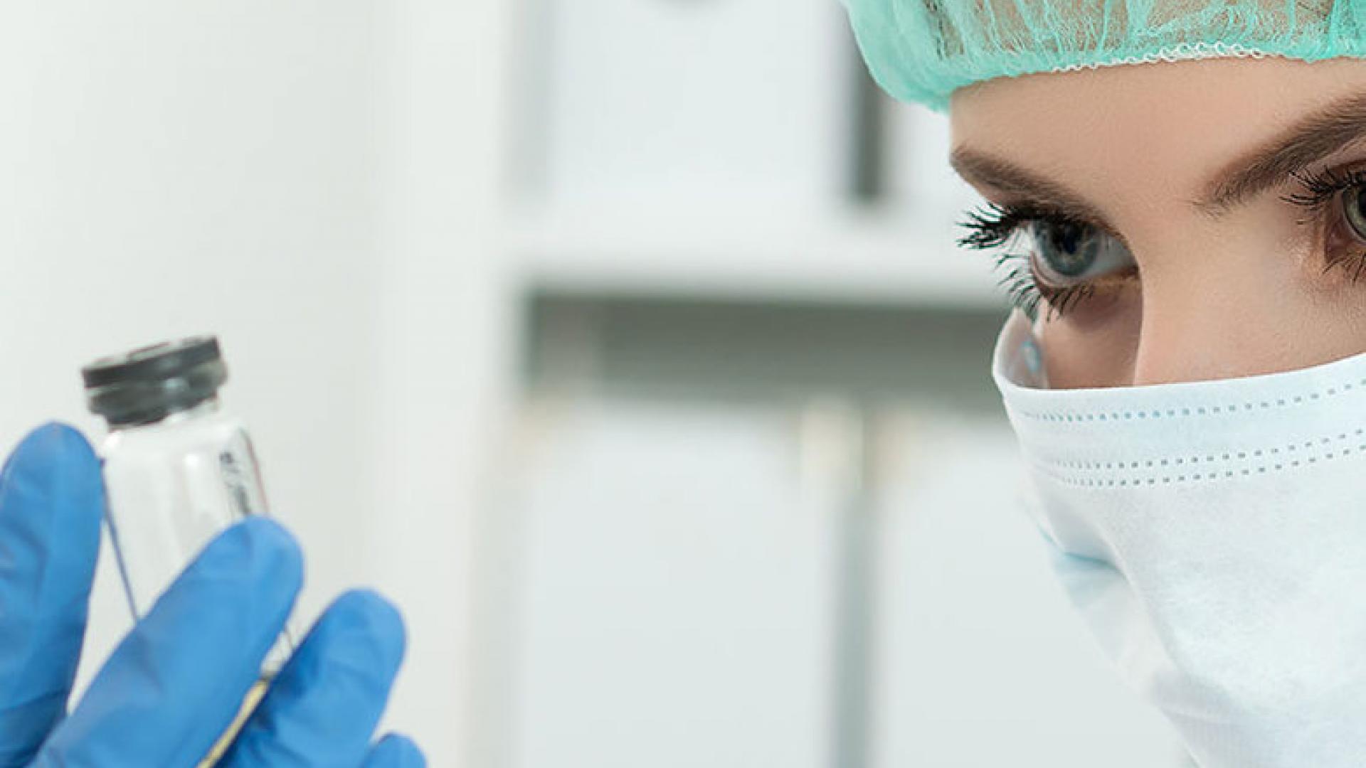 A photograph of someone weaying a medical mask and protective hair covering, viewed from chin to eyebrows, their fingertips covered with a blue latex glove holding a clear glass vial in the lower left corner of the image.