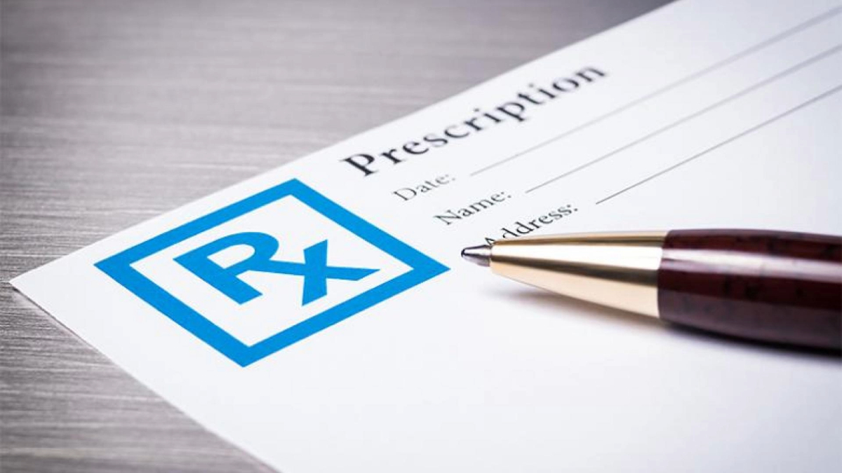 A close-up image of an unfilled medical prescription form with the prominent "Rx" symbol, placed on a wooden surface alongside a brown pen.