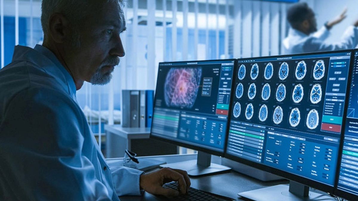 A medical professional reviews brain scans on multiple monitors in a dimly-lit office.