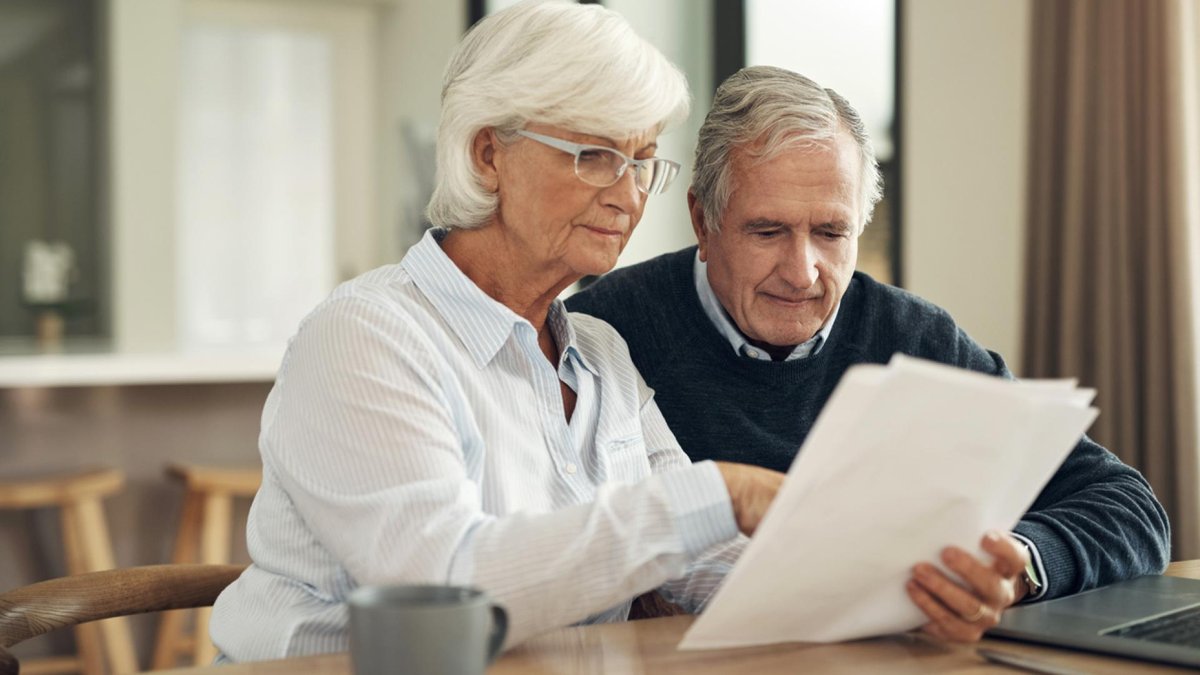 Senior couple sitting together at a table, reviewing paperwork with focused expressions, suggesting they are managing finances or important documents.