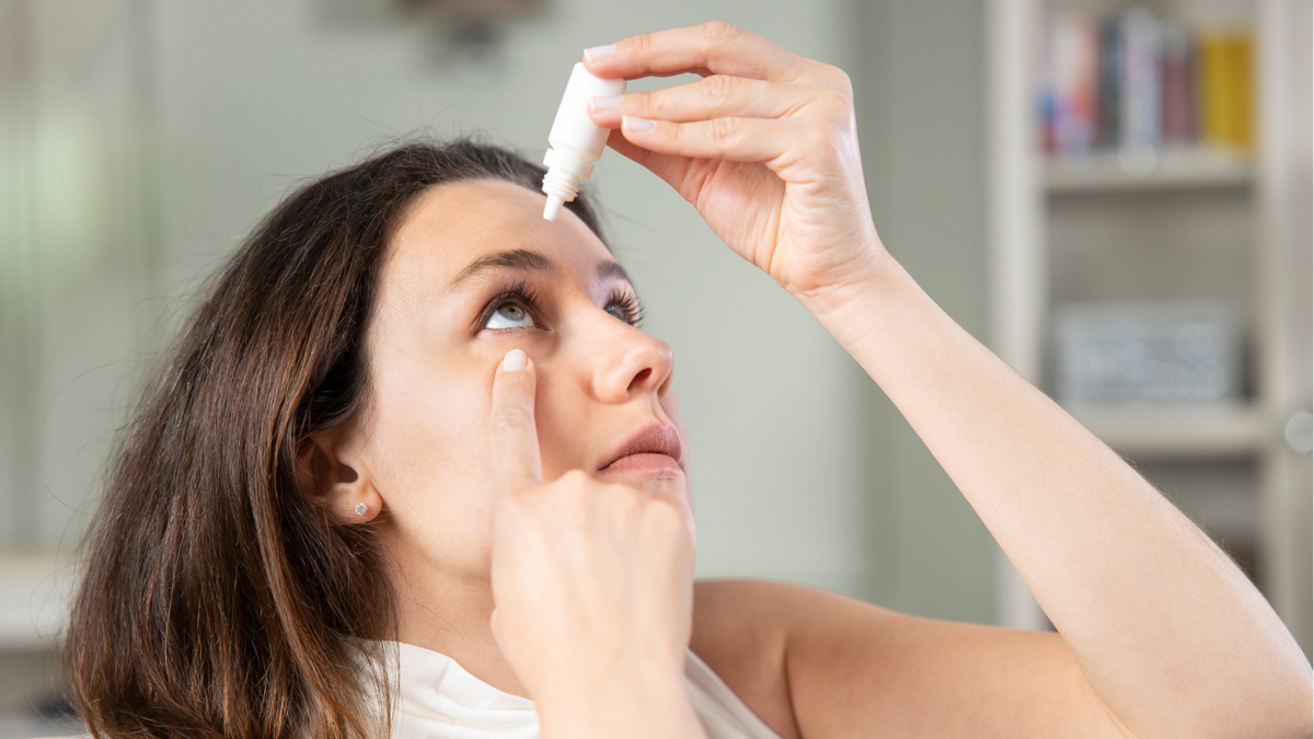 A woman sitting indoors, holding an eye drop bottle above her eye, about to apply the drops with her finger gently pulling down her lower eyelid.