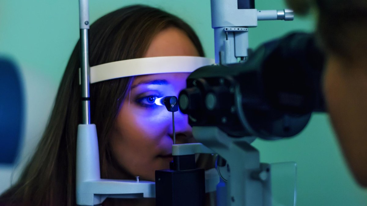 A woman is seated in front of an optical device, having her eyes examined under a blue light, possibly for glaucoma or another eye condition.