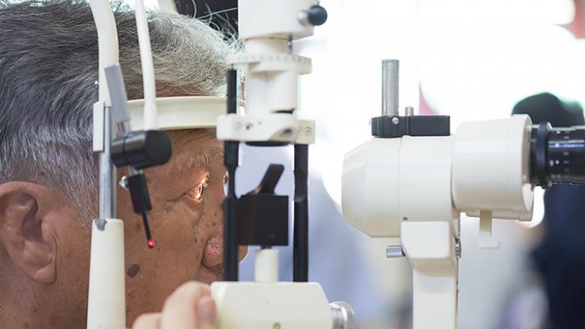 An elderly man is seated in front of an optical device, having his eyes examined by a slit lamp, a common instrument used in optometry.