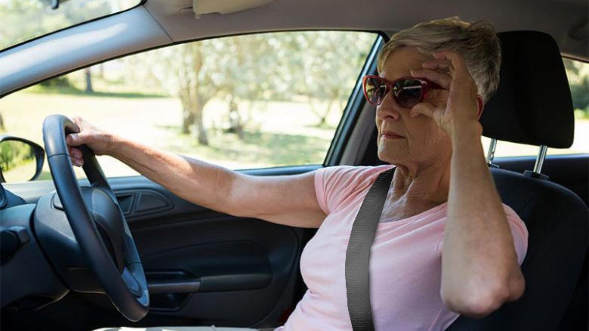 An older woman wearing sunglasses and a pink shirt is driving a car. She holds the steering wheel with one hand and has the other hand raised to her head, appearing to shield her eyes from the sun or focus her vision. She is wearing a seatbelt and appears to be concentrating.