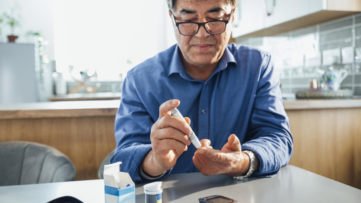 A man in a blue shirt is sitting at a table in a kitchen, using a small device to check his blood sugar levels by pricking his finger. A glucose monitor and test strips are also visible on the table.