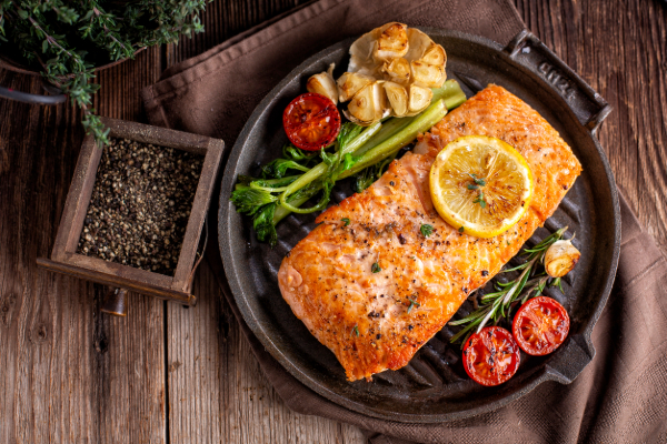 Grilled salmon fillet garnished with lemon slices and herbs, served with asparagus and cherry tomatoes on a rustic wooden table.