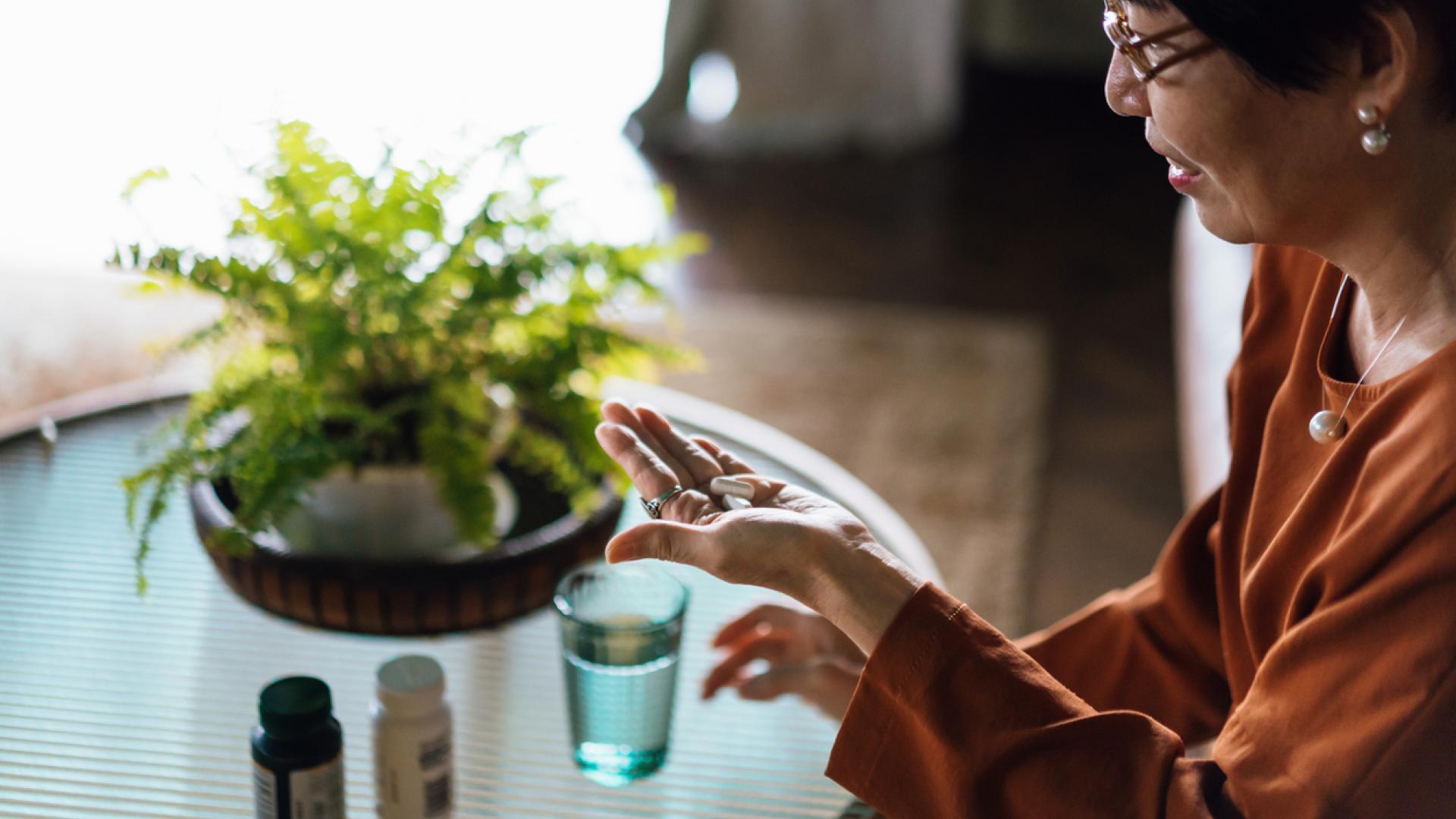 an older woman, viewed from the side and slightly above, holds two pills in her open palm. In the background, a glass of water and a plant are on a glass table