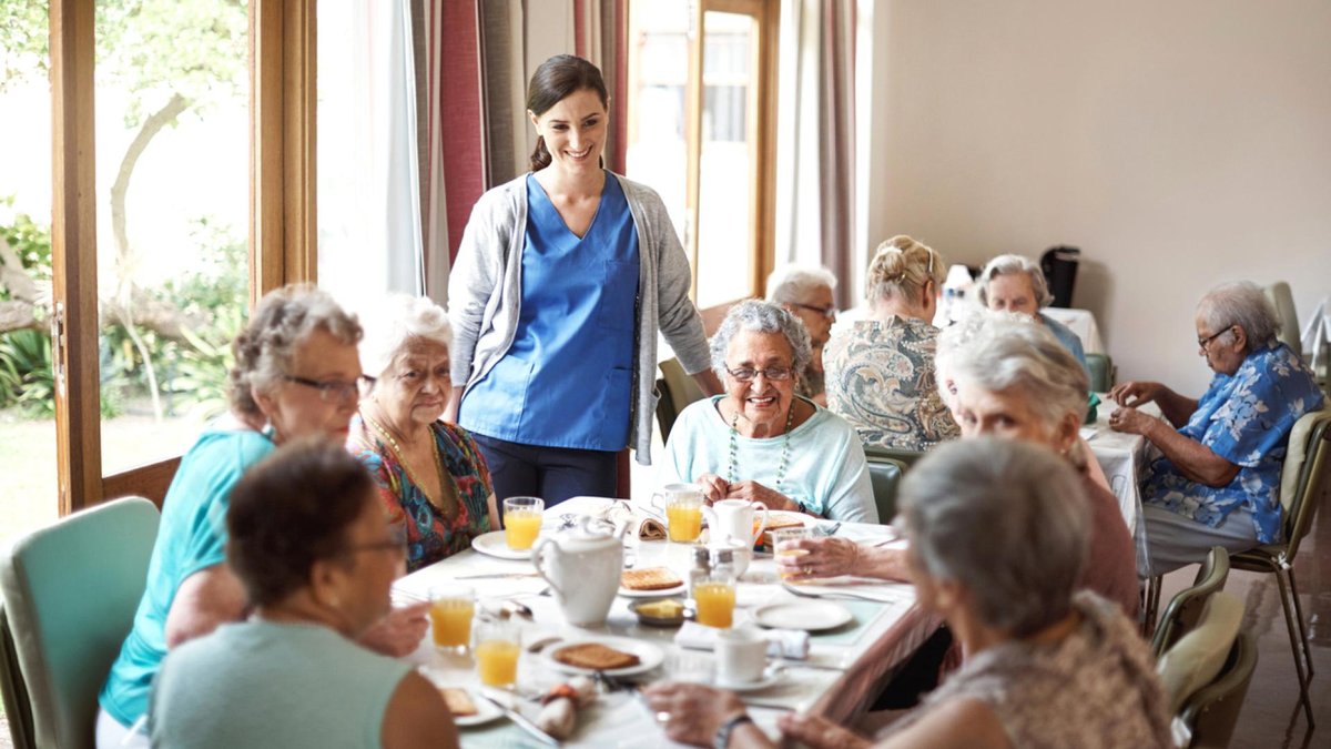 A group of senior women gathered around a table enjoying breakfast together at a care facility, with a nurse standing nearby, smiling.