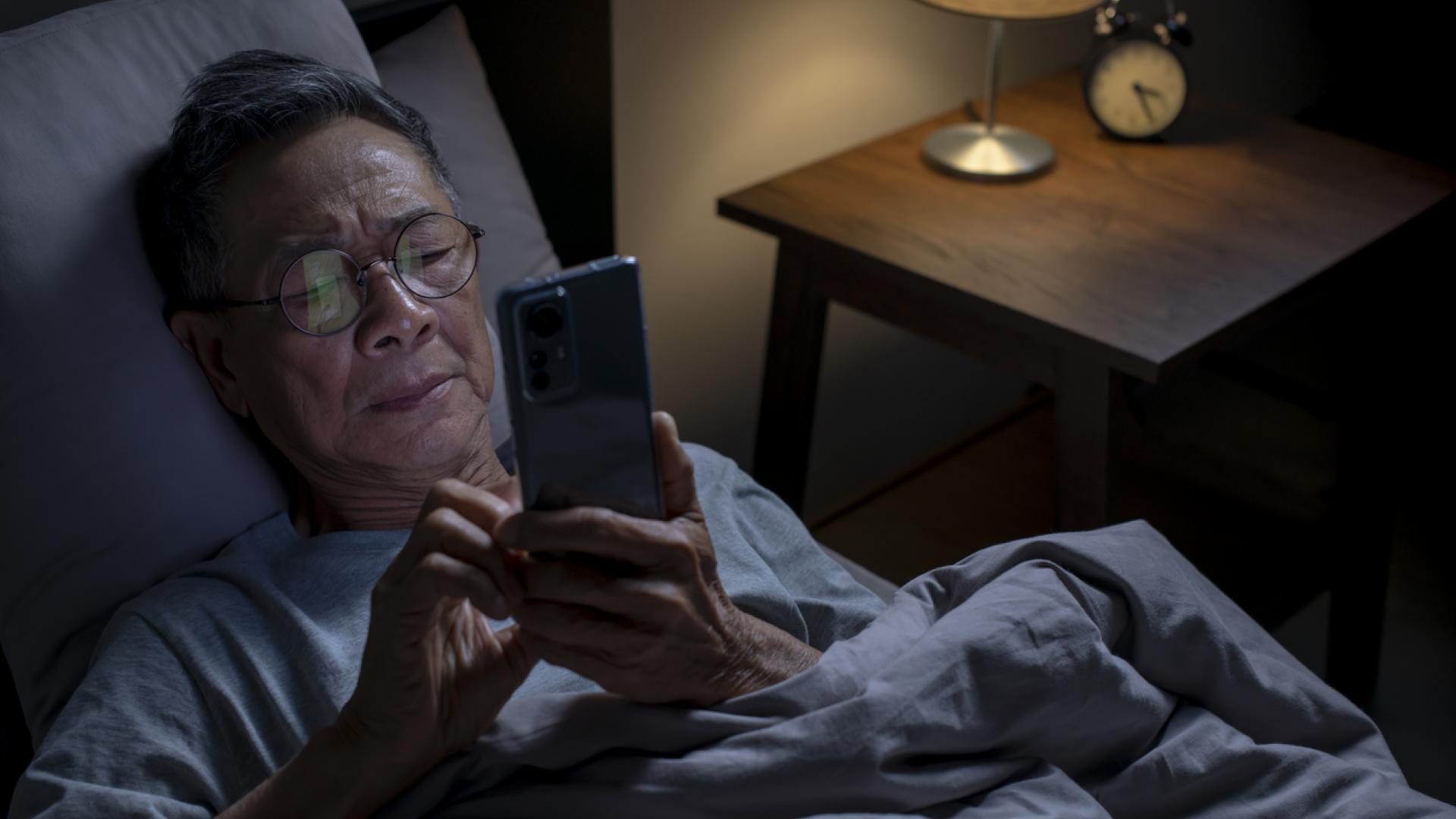 A senior man lying in bed at night, wearing glasses and looking at a smartphone. The dimly lit room features a bedside table with a lamp and an alarm clock, suggesting he may be experiencing difficulty sleeping or insomnia.