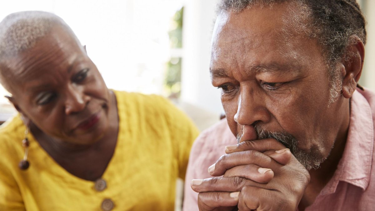 A concerned elderly woman in a yellow shirt sits beside an older man who appears upset, with his hands clasped and resting against his face.