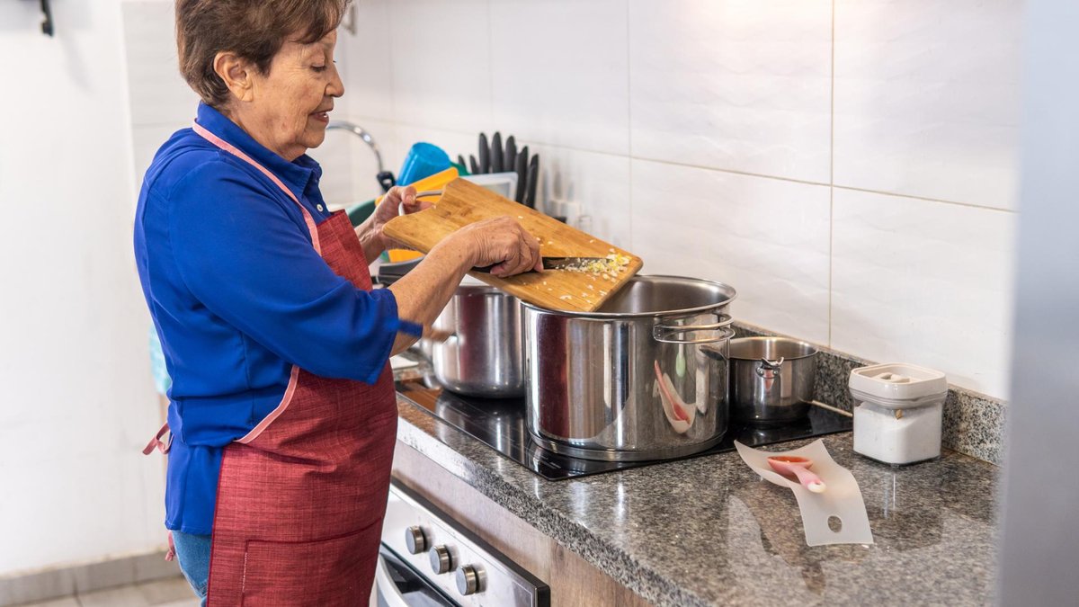 An elderly woman wearing a red apron is cooking in her kitchen, stirring ingredients into a large aluminum pot on the stove, surrounded by other kitchenware.