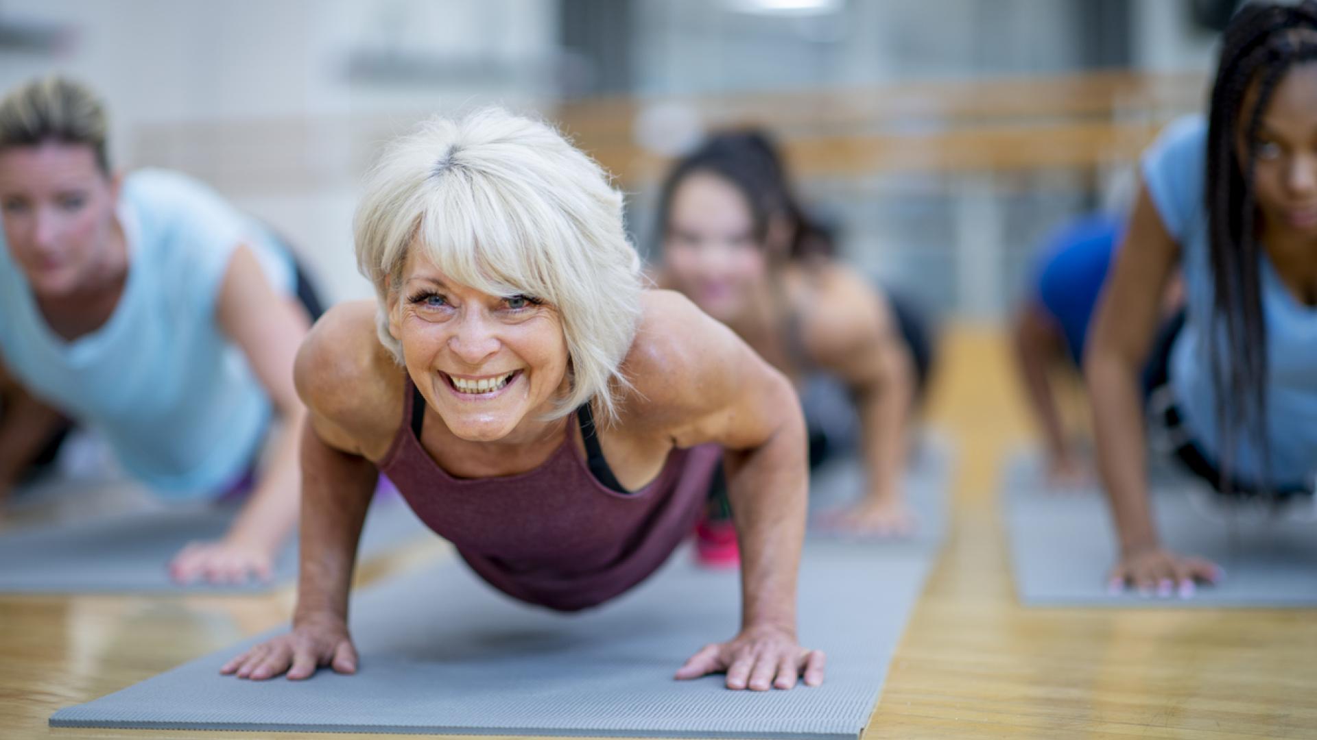 A photo of a senior woman in push-up position in a yoga mat with several younger, out-of-focus people in the photo as well.