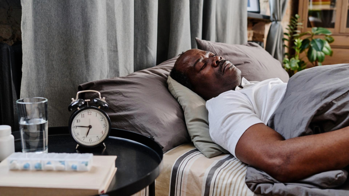 Person sleeping peacefully on a bed with a pillow, white bedsheets, and a gray blanket. Beside the bed is a nightstand with an alarm clock showing the time, a glass of water, and a strip of medication tablets.