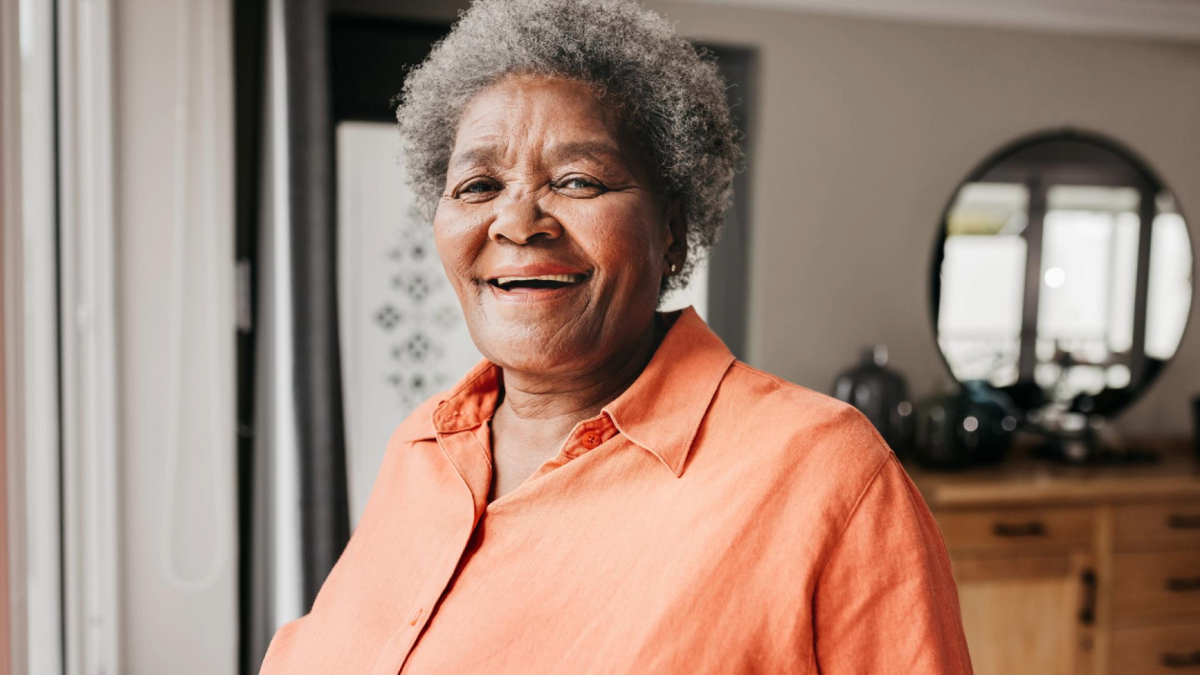 A smiling person with gray hair, wearing an orange shirt, standing in a modern kitchen.