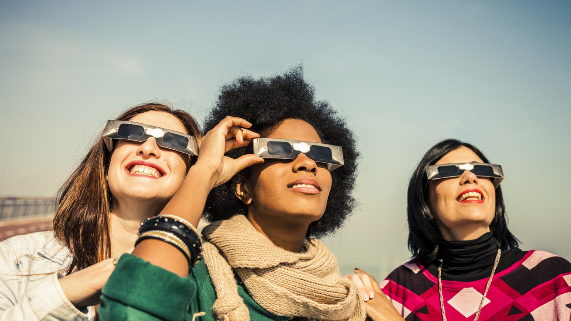 three women wearing solar eclipse protection glasses stare at the sky, smiling