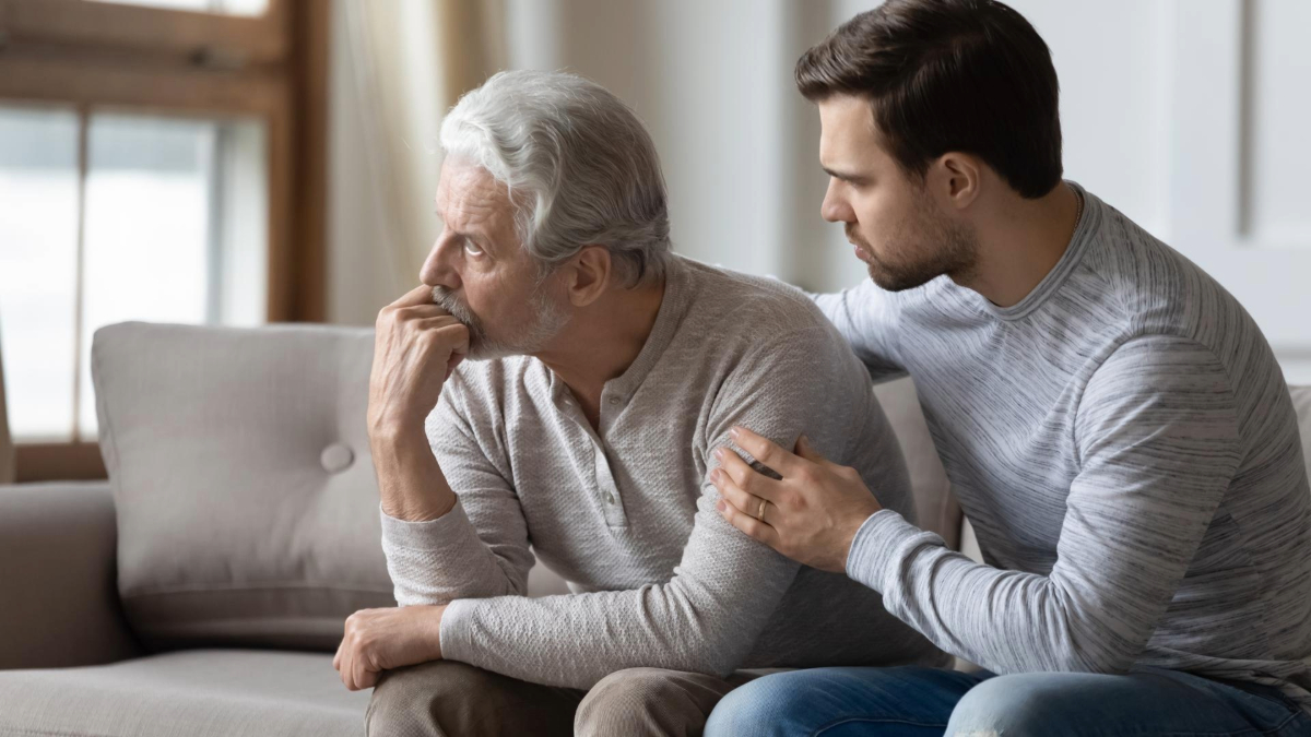 An older adult and a younger adult sitting together on a sofa, with the younger adult comforting the older by placing a hand on their shoulder. Both are looking thoughtfully to the side.