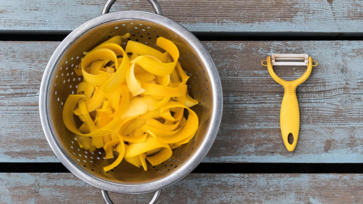 A stainless steel colander filled with squash ribbons on a wooden table, next to a yellow-handled peeler.